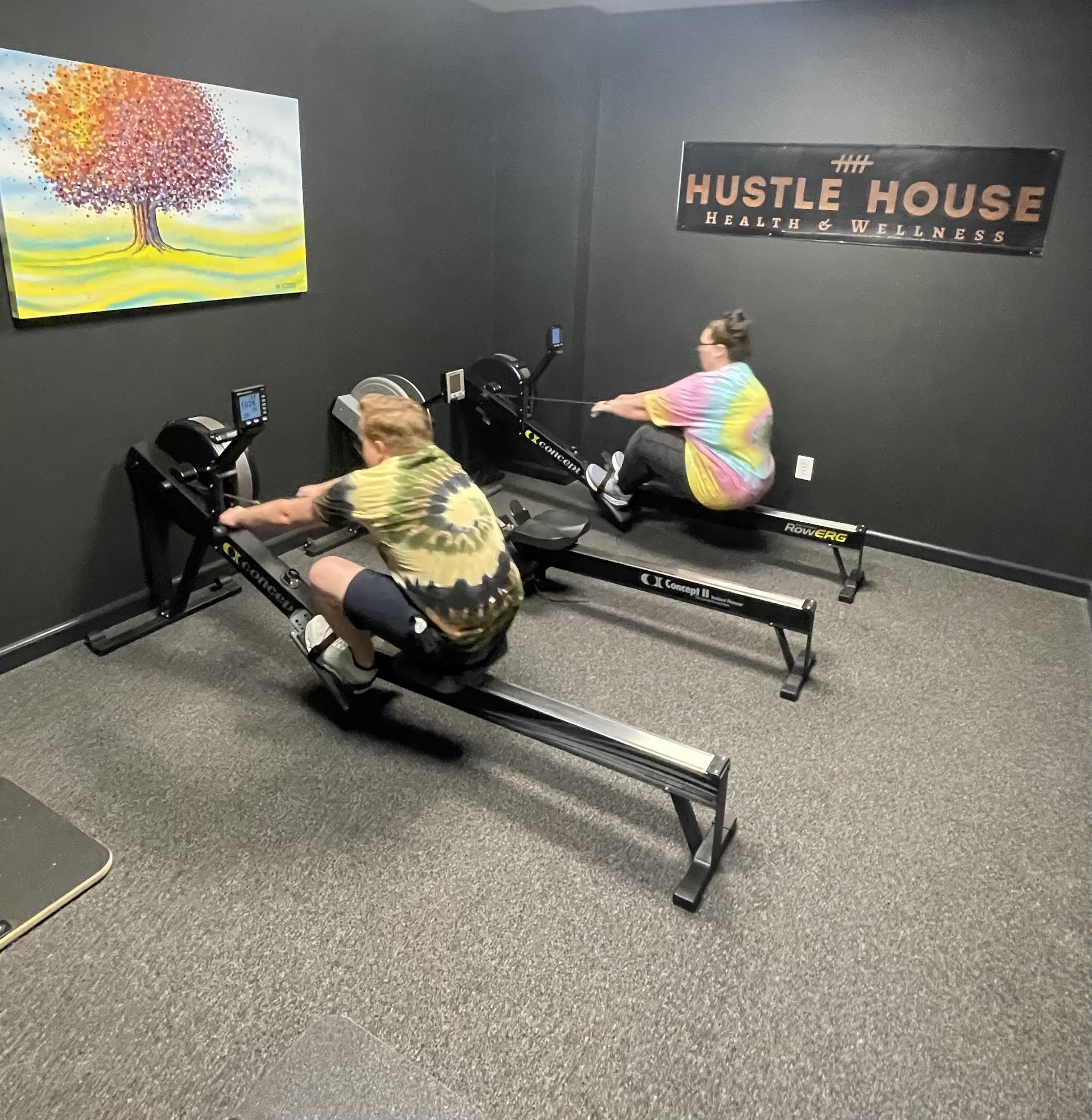 Two members rowing side by side beneath the studio sign at Hustle House Health & Wellness in Woodstock, GA